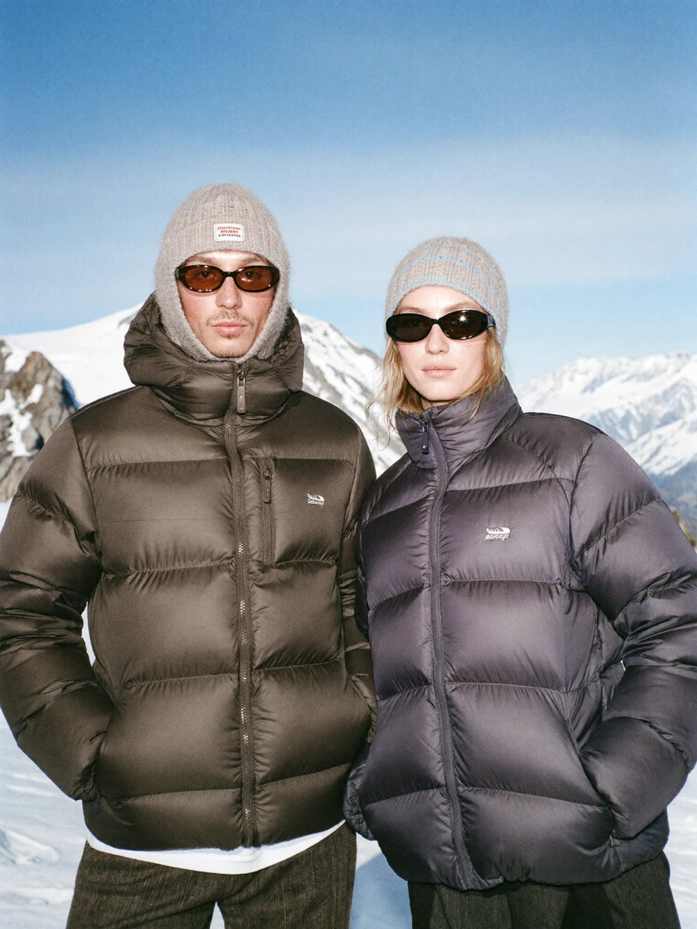 Two models in dark puffer jackets against a snowy mountain landscape