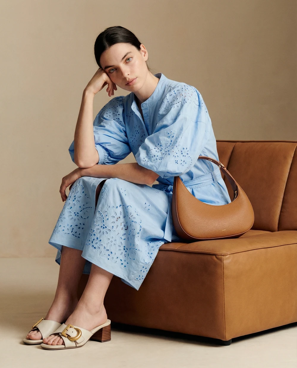 Woman in an embroidered blue dress sitting on a brown leather sofa with a tan handbag