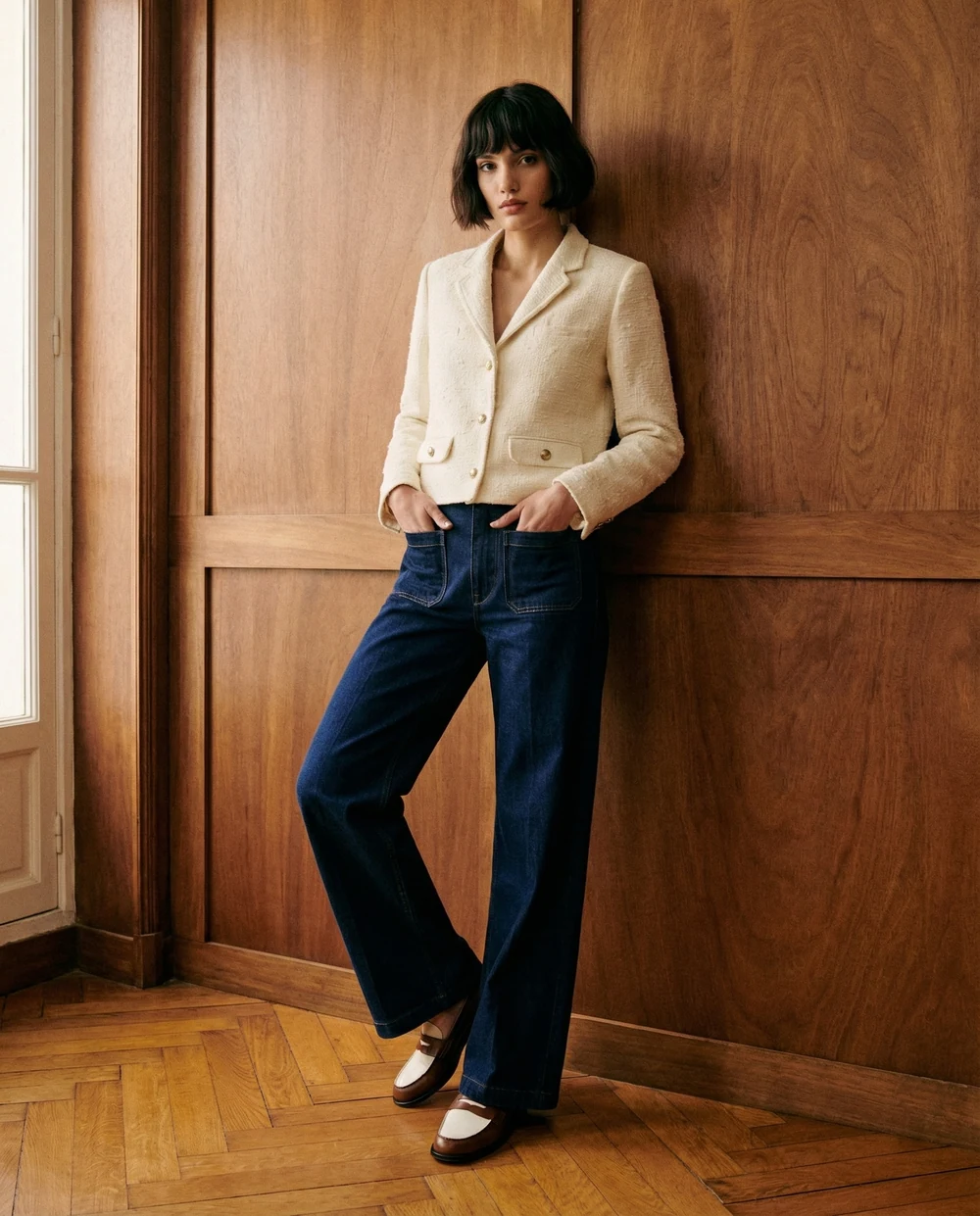 Woman in a cream jacket and dark denim leaning against a wood panelled wall